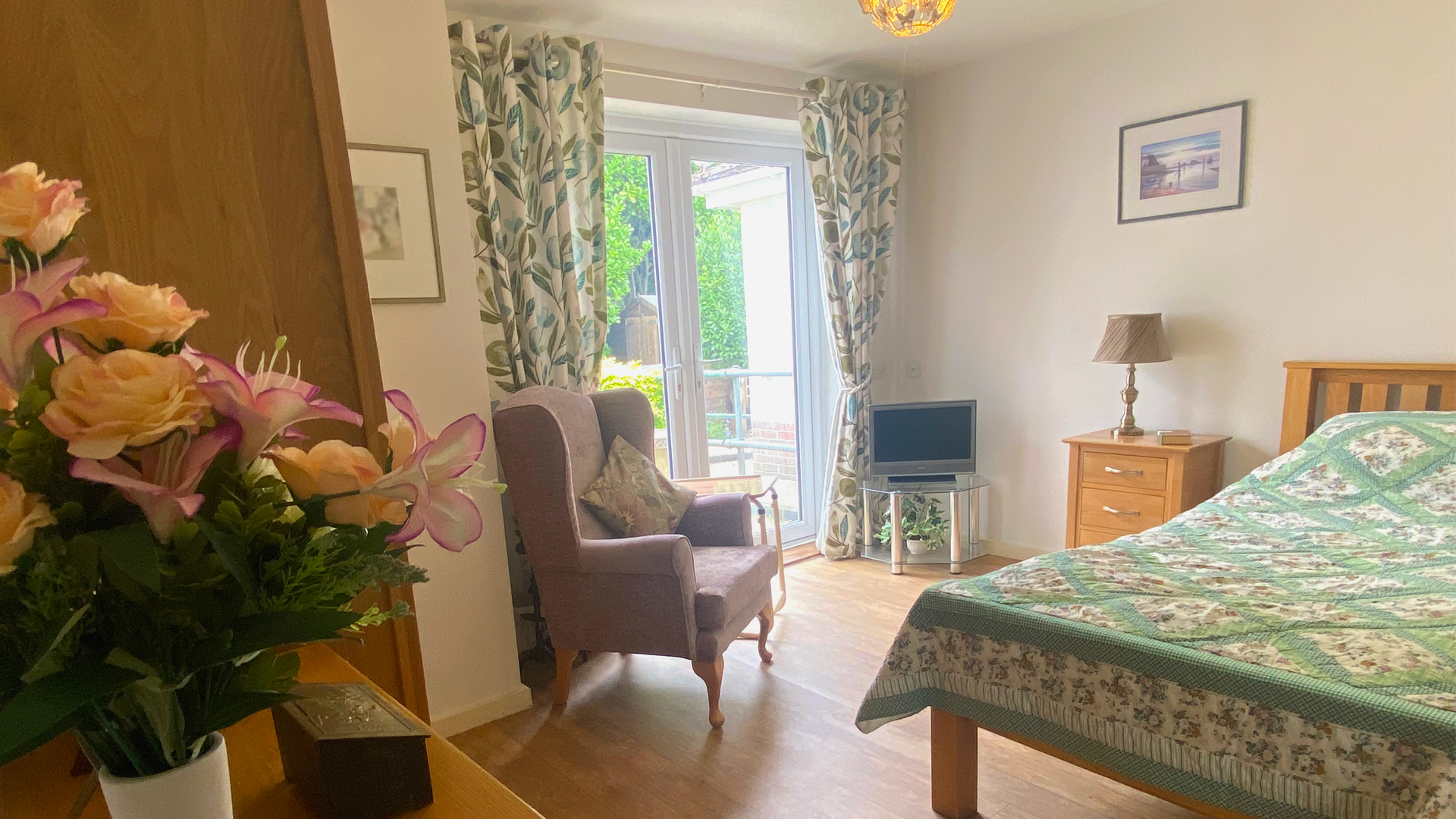 A bright and cosy bedroom at the care home, with floral bedding, comfy armchair, bedside table and lamp, wardrobe and chest of drawers and a big sunny window looking out onto the garden, decorated with flowers and paintings