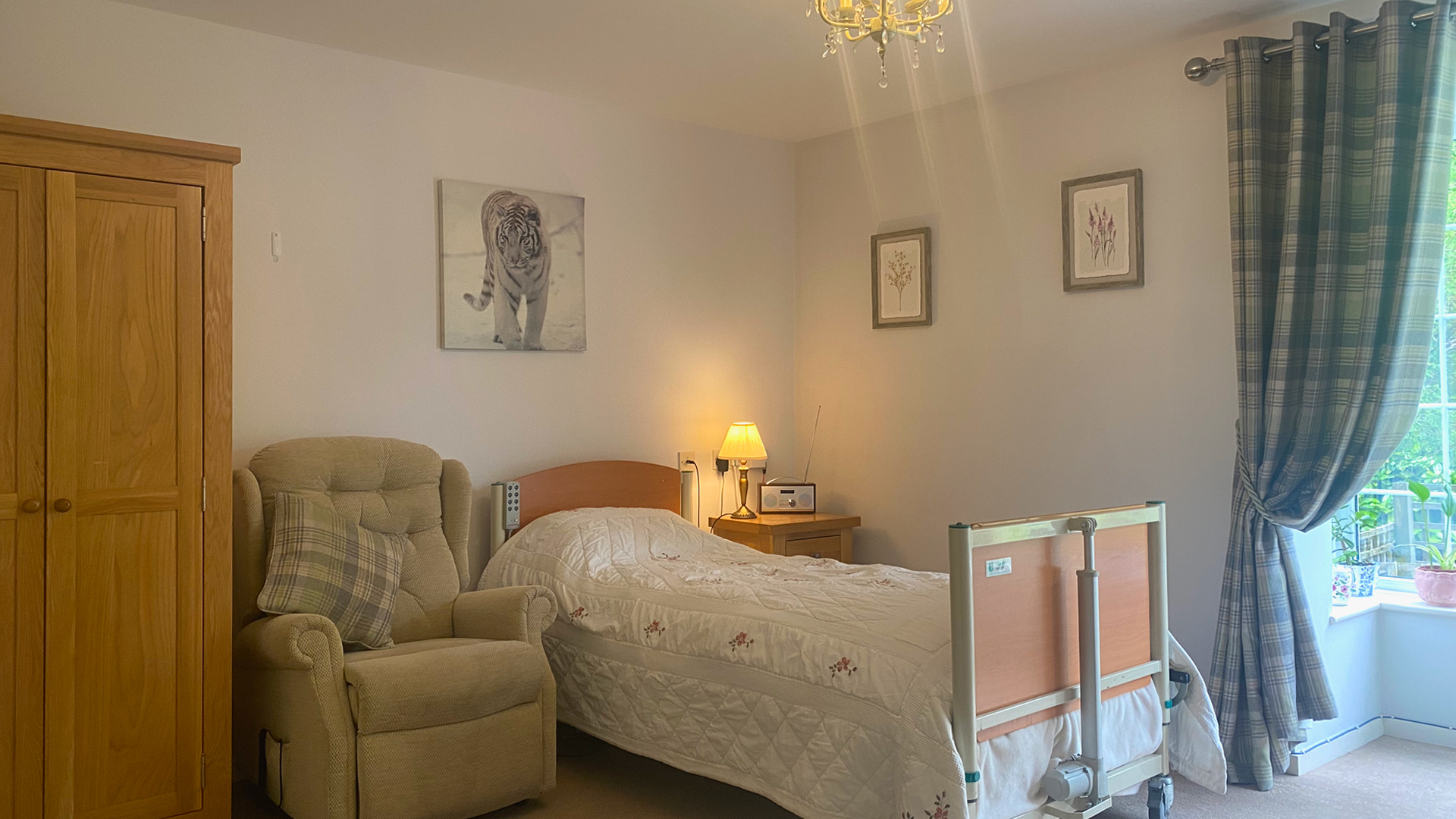 A homely looking bedroom at the care home, with a tidy bed, wardrobe, reclining chair, sunny window and decorated with paintings and flowers