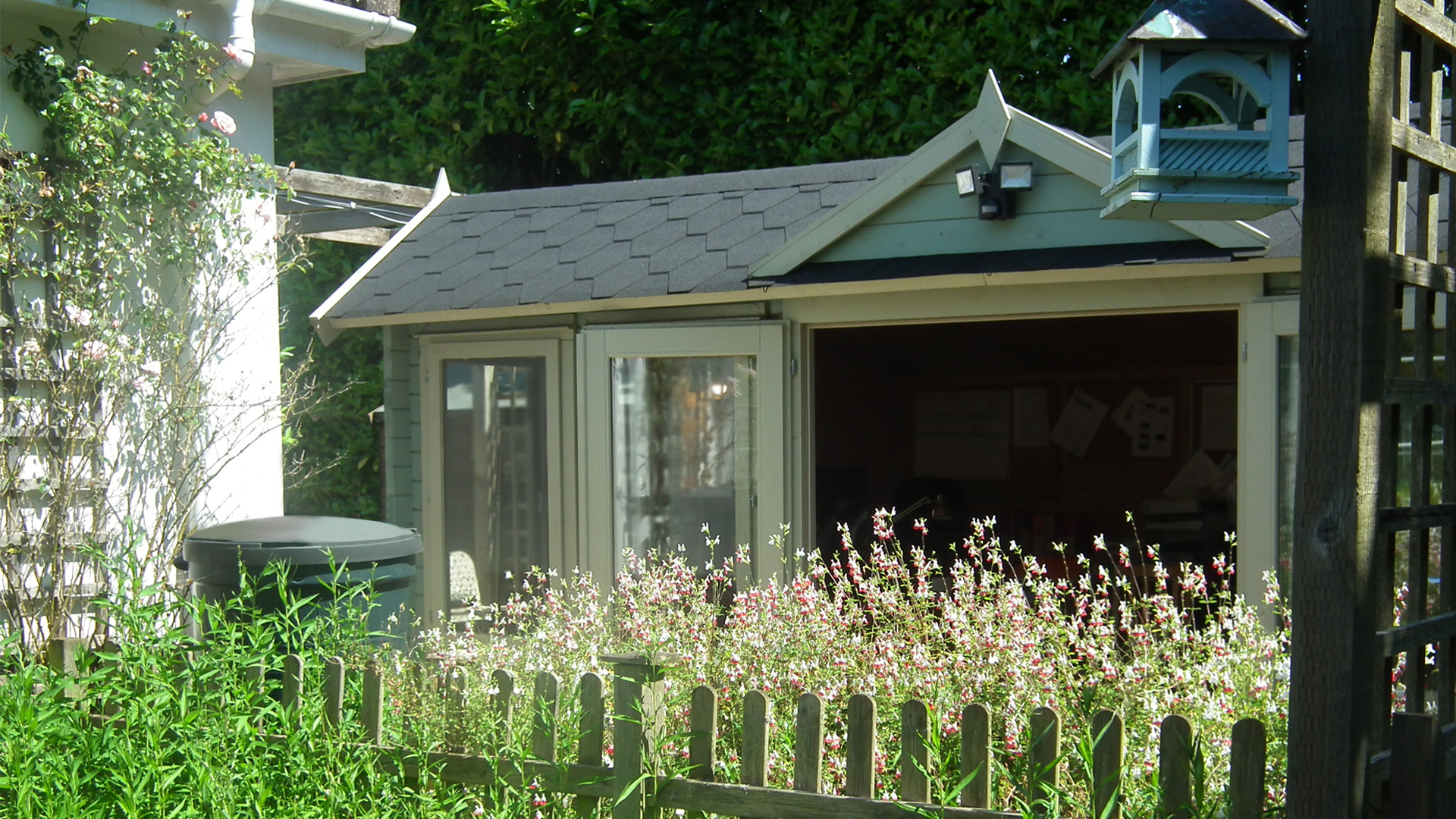 The care home office cabin photographed behind hot lips flowers, a bird house and a small wooden fence in the care home garden
