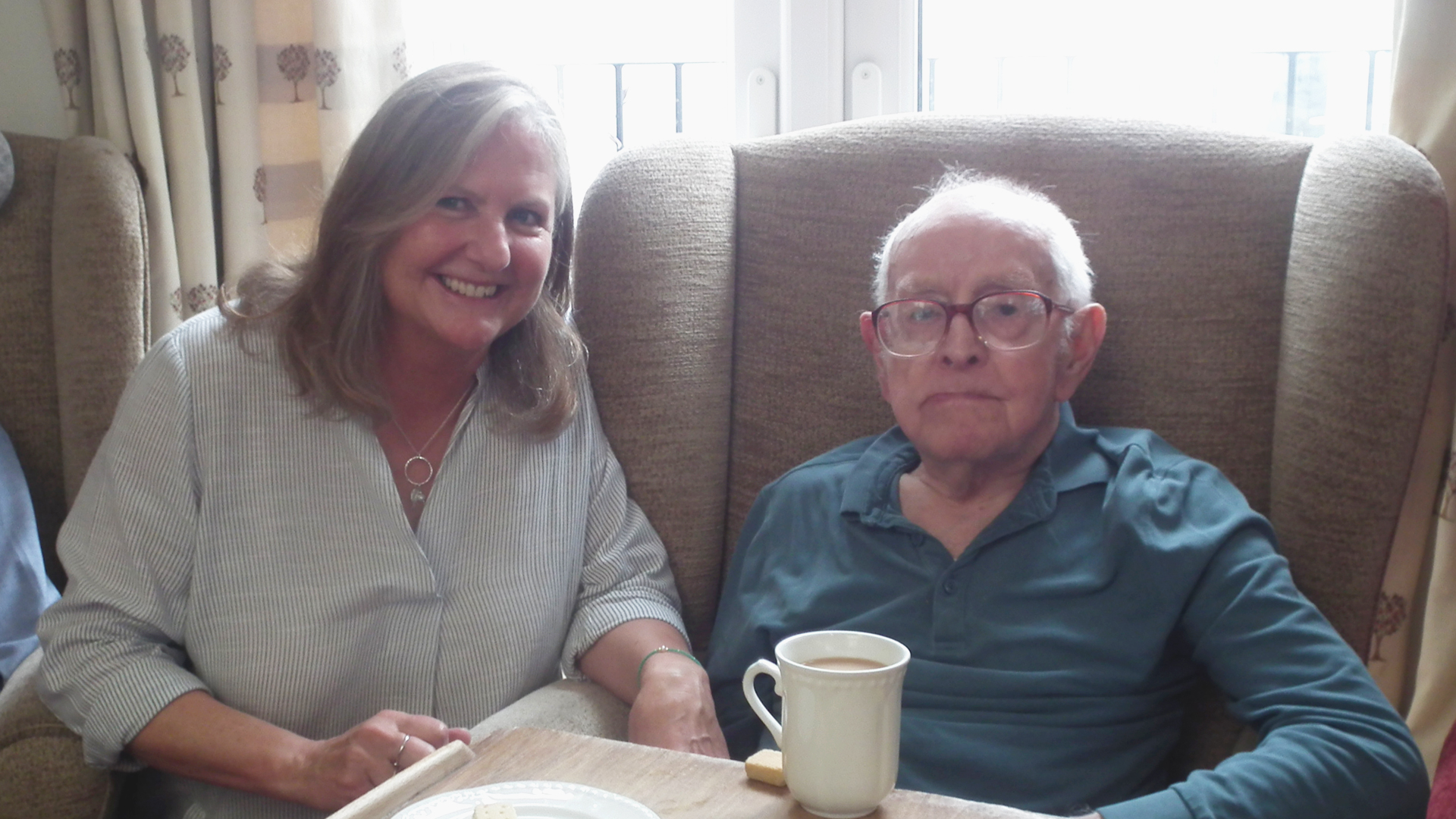Debbie the Care Staff Manager smiles while holding the hand of a resident sat in an armchair with a cup of tea and biscuit
