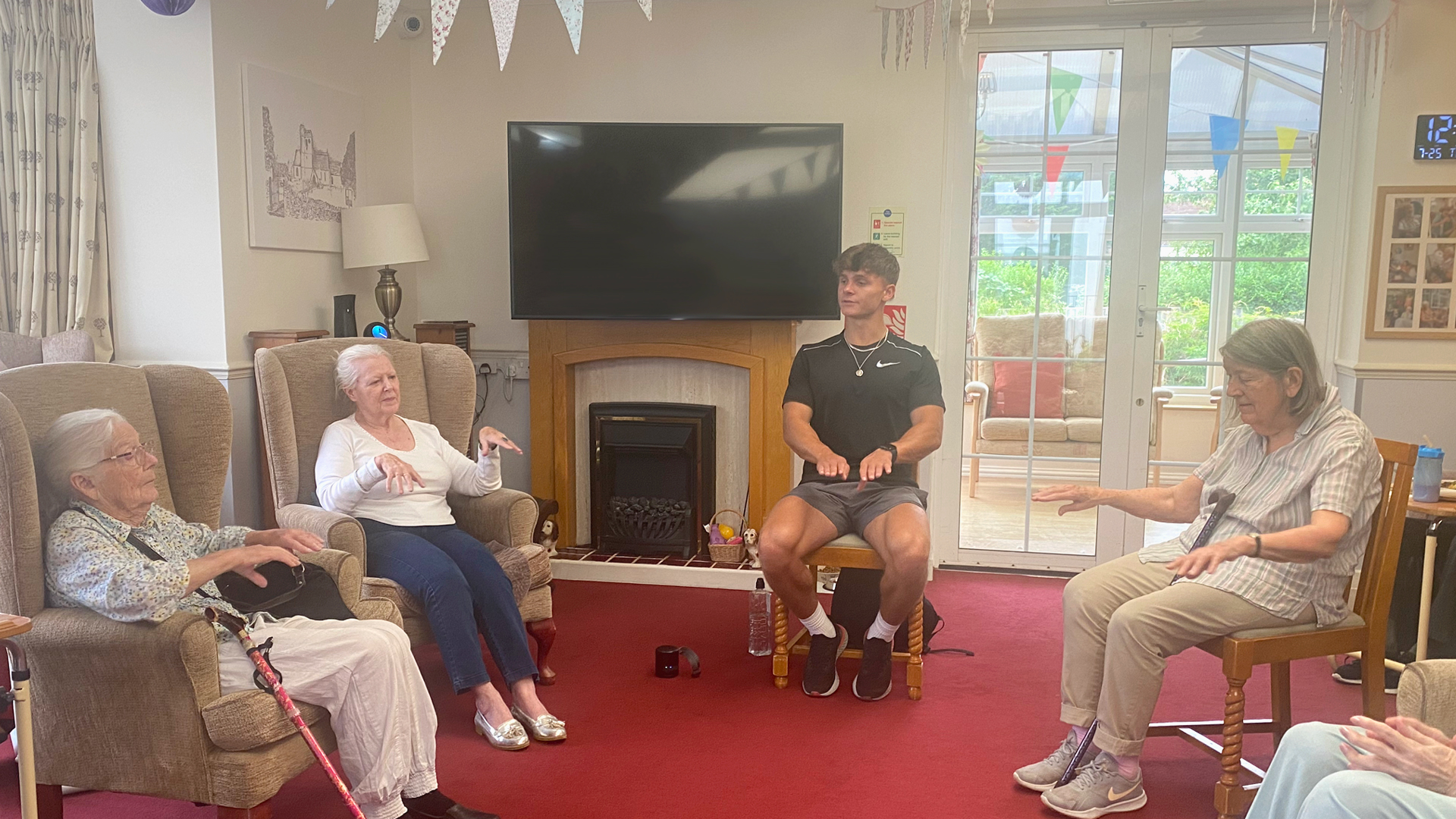 A fitness instructor sits in a circle with some of the care home residents, everyone makes swimming motions with their hands