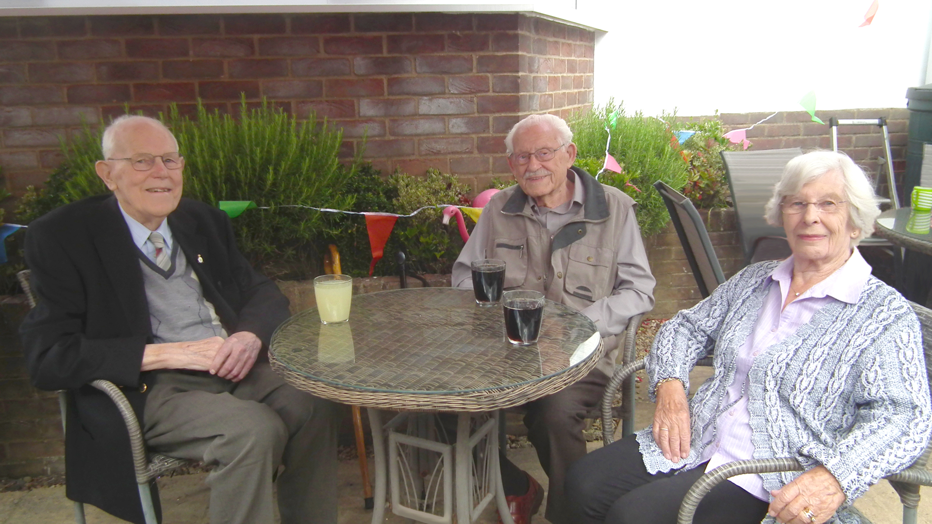 A care home resident sits with two visitors at the summer party, sat outside in the decorated garden with glasses of squash