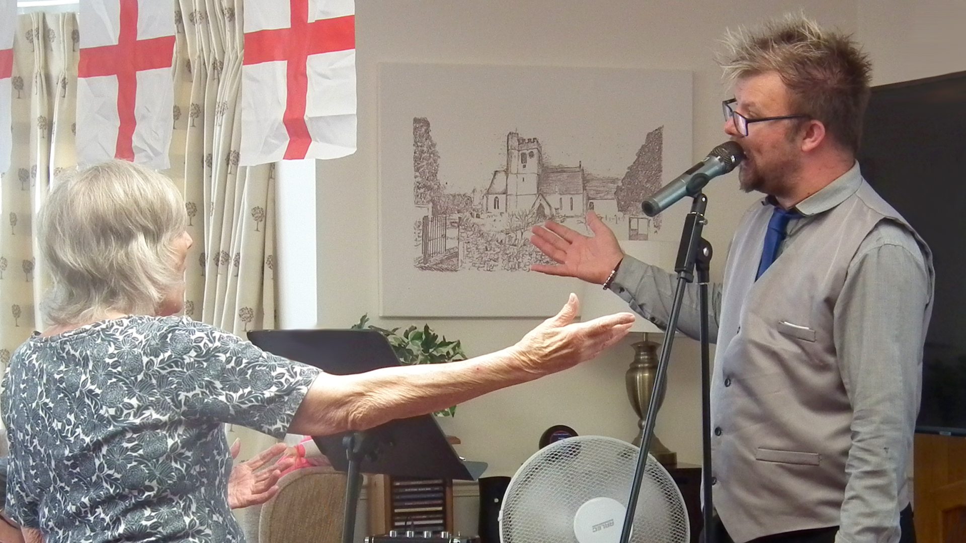 A care home resident stands and sings with her arms out as the visiting entertainer sings a song with her