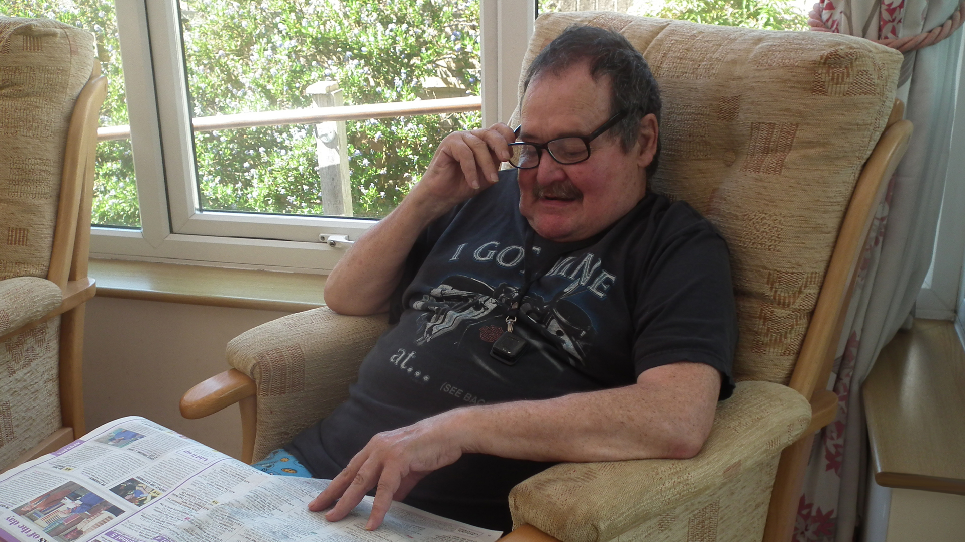 A care home resident holds his reading glasses to his face as he reads the newspaper in the sunny conservatory