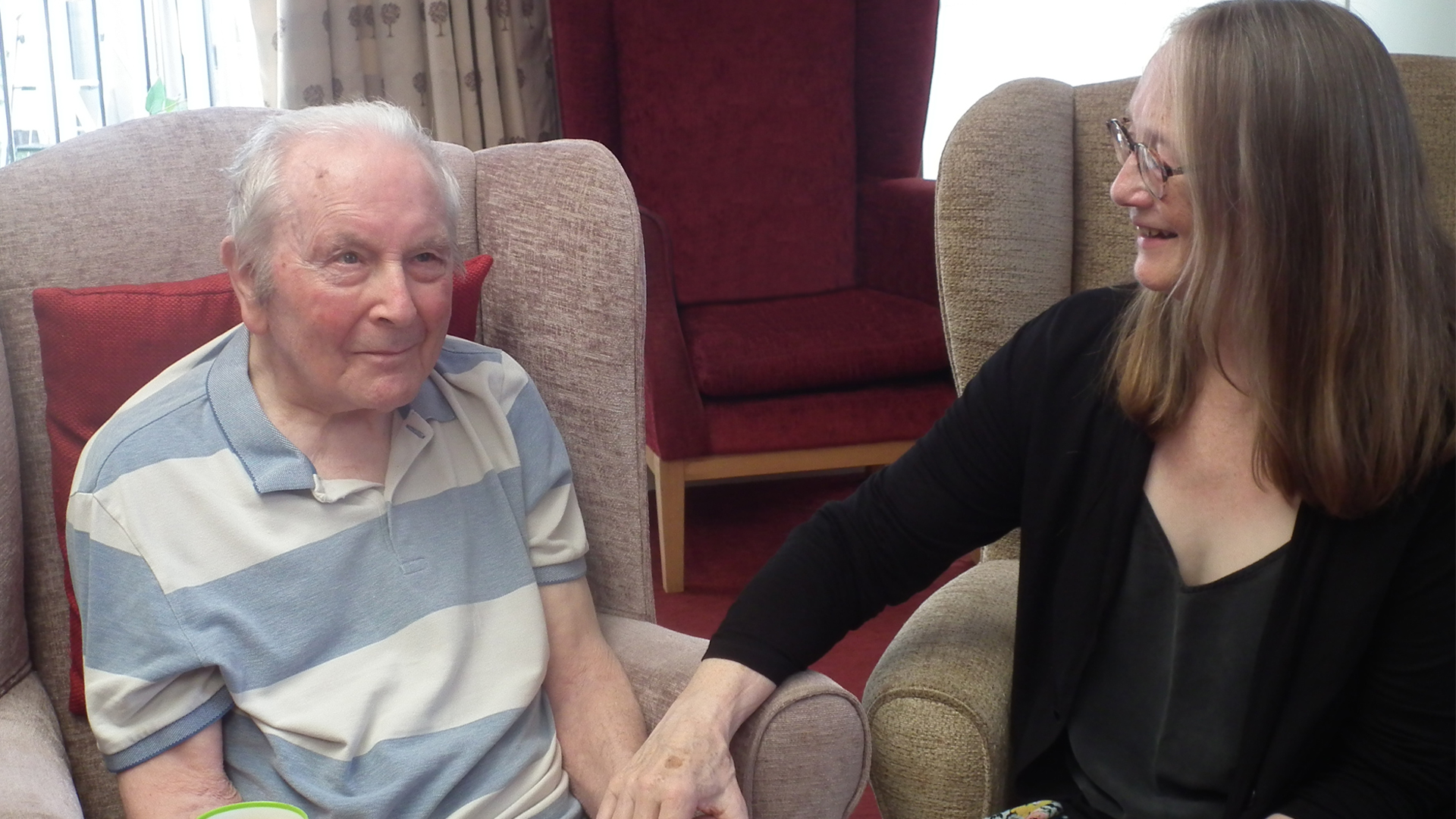 A resident at the care home sits in an arm chair, smiling and holding hands with his daughter