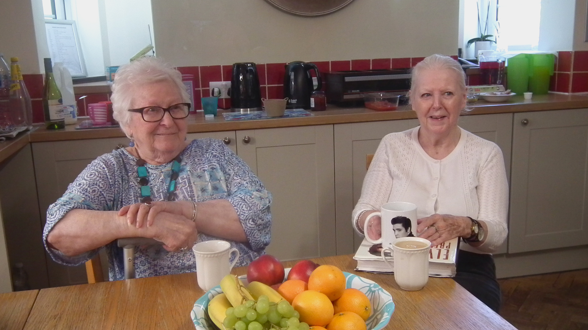 Two care home residents sit at a table with a fruit bowl in the kitchen, both have a cup of tea and smile