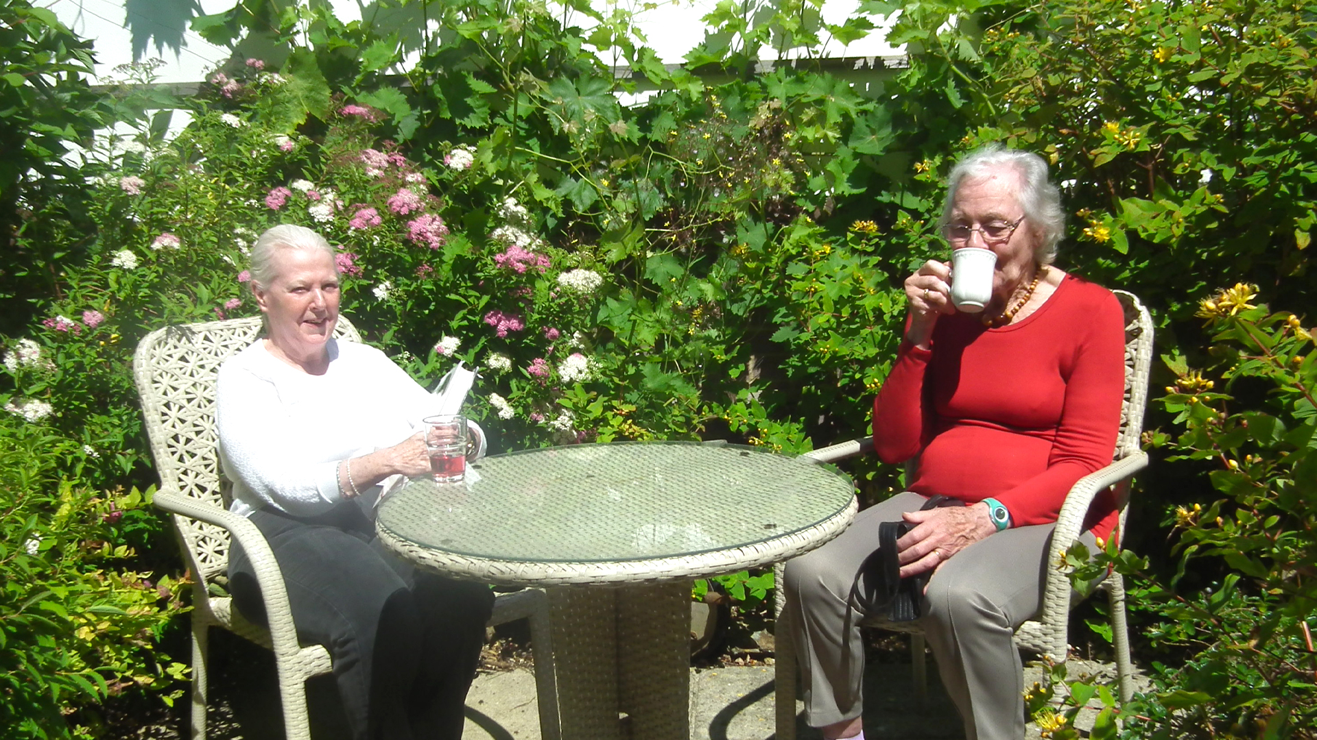 Two care home residents sit outside in the sunny garden, one is reading a book with a glass of squash while the other sips her cup of tea