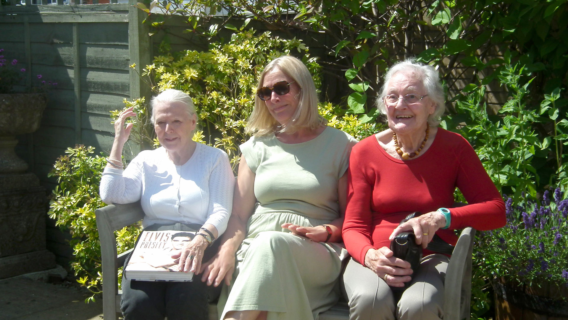 Two care home residents sit beside one of the staff members on a bench in the garden, all are smiling