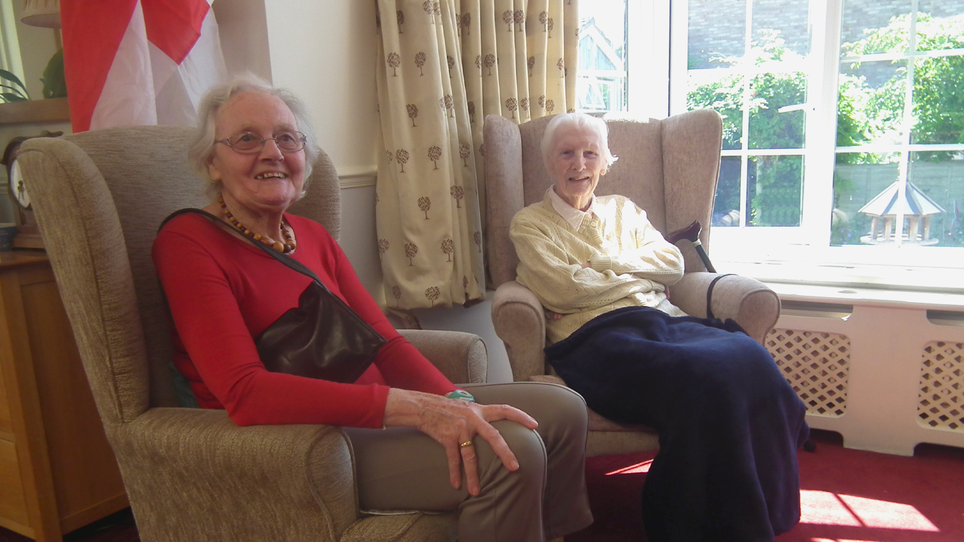 Two care home residents smile while sitting in armchairs in the window of the living room