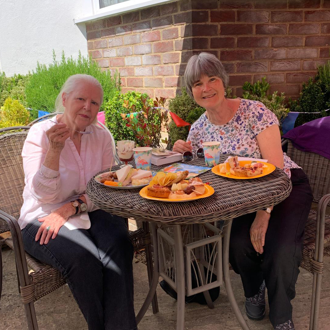 A resident at the care home sits next to her daughter at the summer party. Both are smiling while eating party snacks on bright coloured party plates