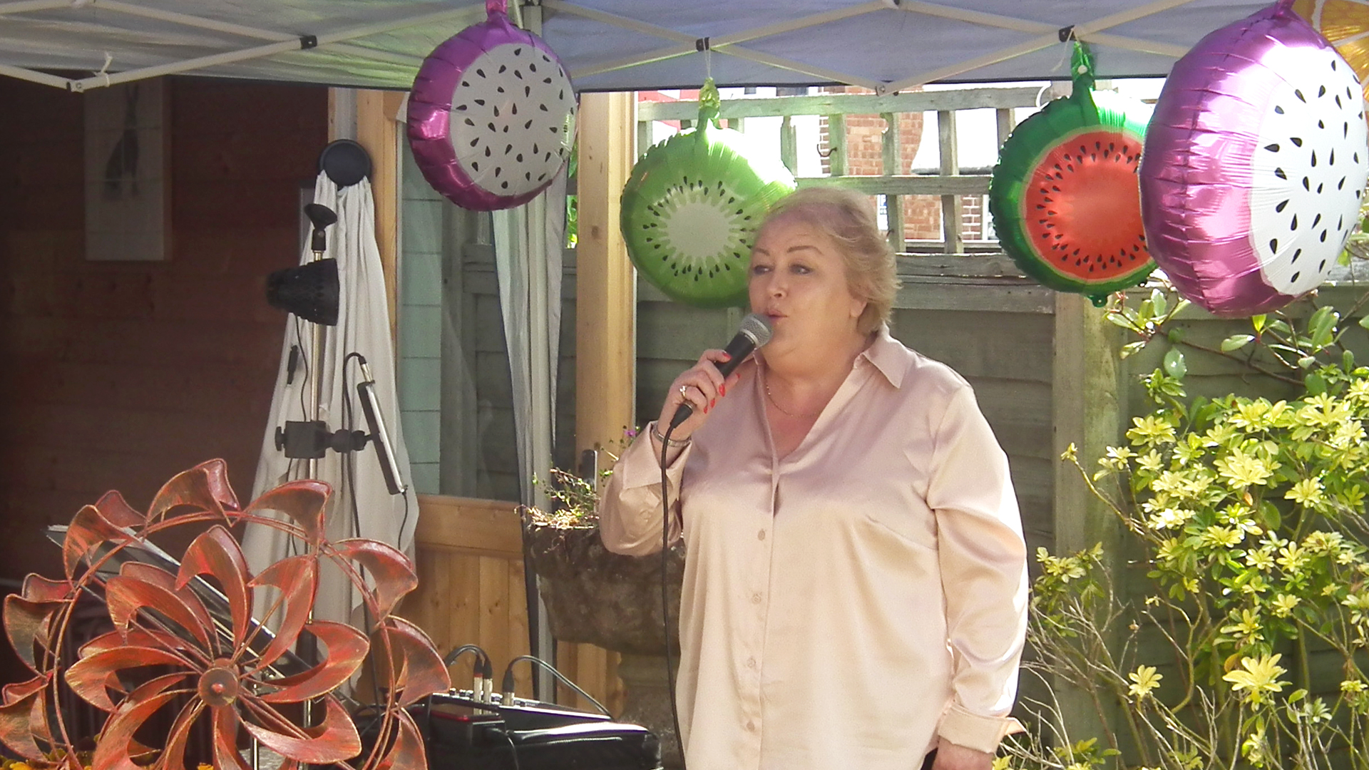 Jackie the singer holds the microphone while singing to the residents and visitors at the care home summer party, she stands in front of a backdrop of colourful fruit-themed balloons
