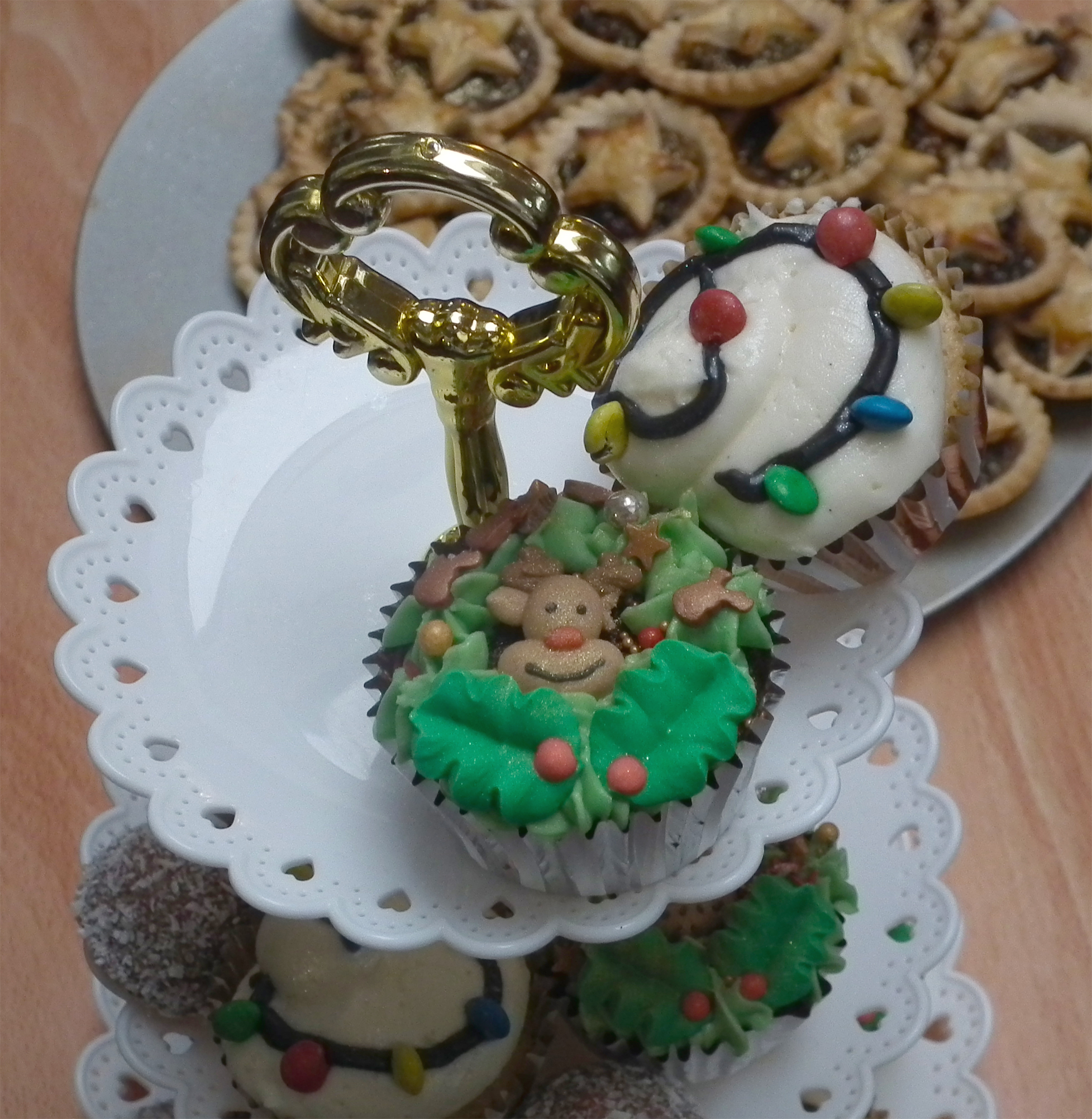 A cupcake stand in front of a plate of mince pies, the cupcakes have Christmas designs on them, one of them with white icing and smarties to look like Christmas lights, the second has green icing and a smiling reindeer cake topper with edible glitter