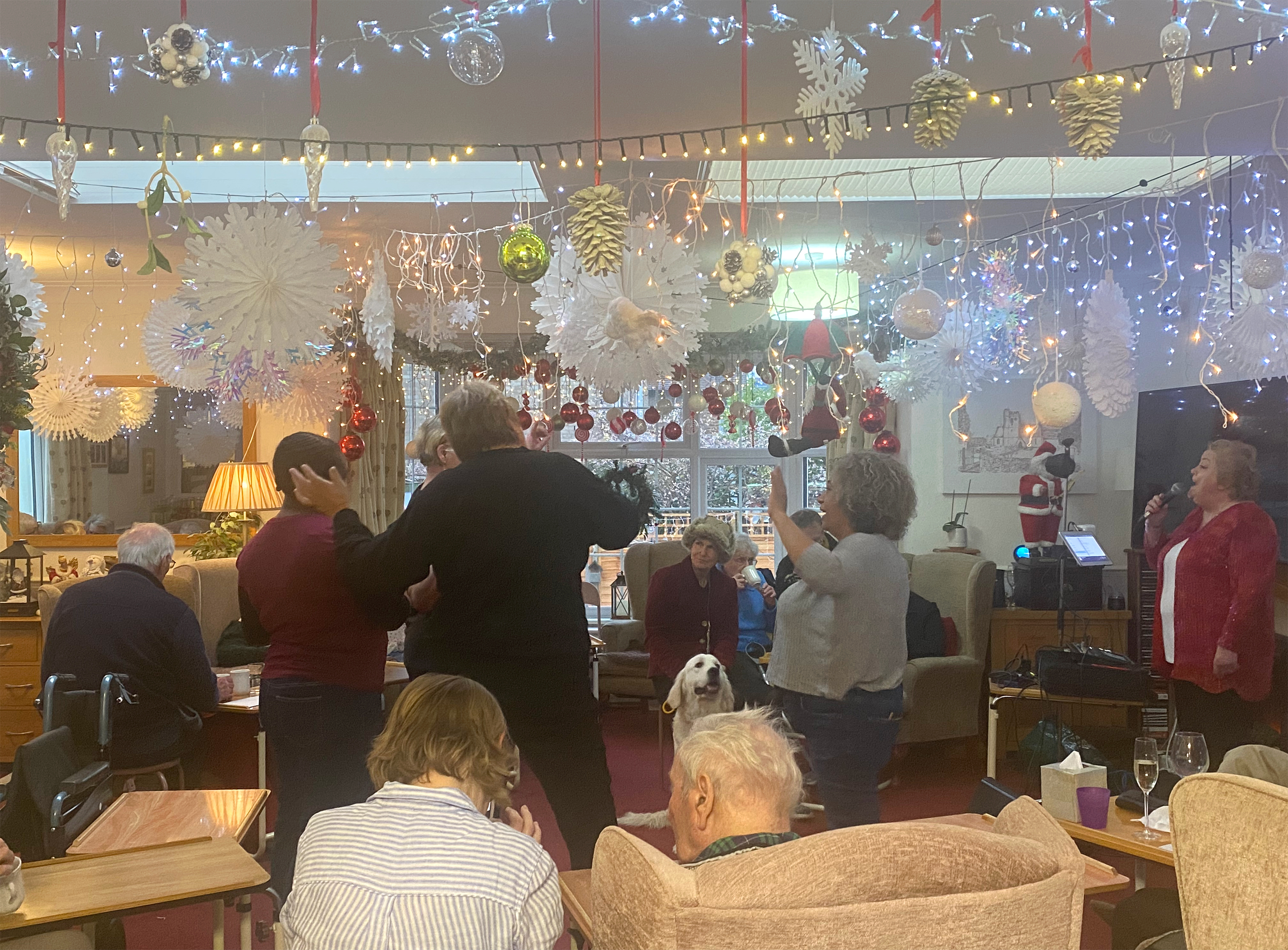 Care staff dancing in the centre of the room while the entertainer sings. The room is decorated with lots of snowflakes, Christmas decorations and lights hanging from the ceiling, and the residents and their families chat while a golden retriever sits in the middle of the room