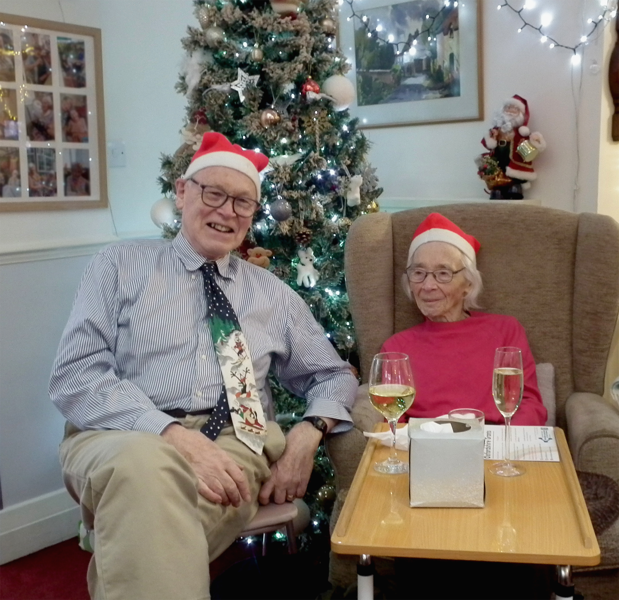 A resident and her relative sit and smile, wearing Christmas hats and with glasses of wine. Behind them is a decorated Christmas tree and Christmas lights