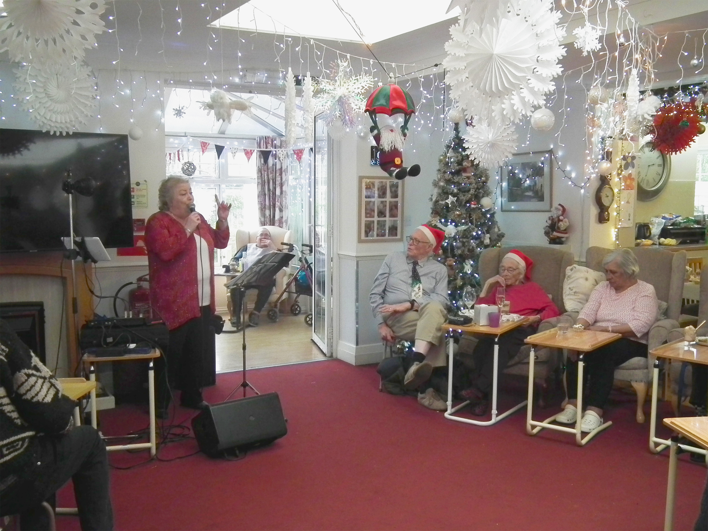 The entertainer sings to the room with residents and their relatives listening. Lights and paper snowflakes fill the ceiling and look very festive