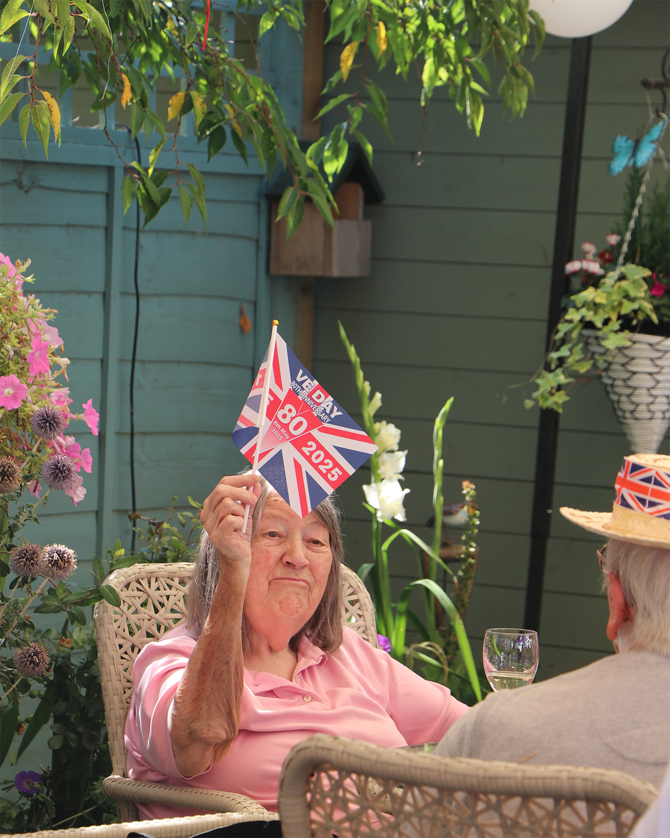 A resident holds their VJ day 80th anniversary flag, she wears a pale pink shirt and sits in front of a garden of colourful flowers