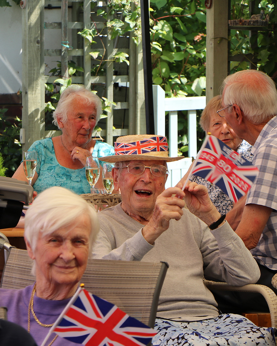 A smiling resident wears a summer hat with the British flag on, and waves a VJ day 80th anniversary flag, people sit and chat in the background