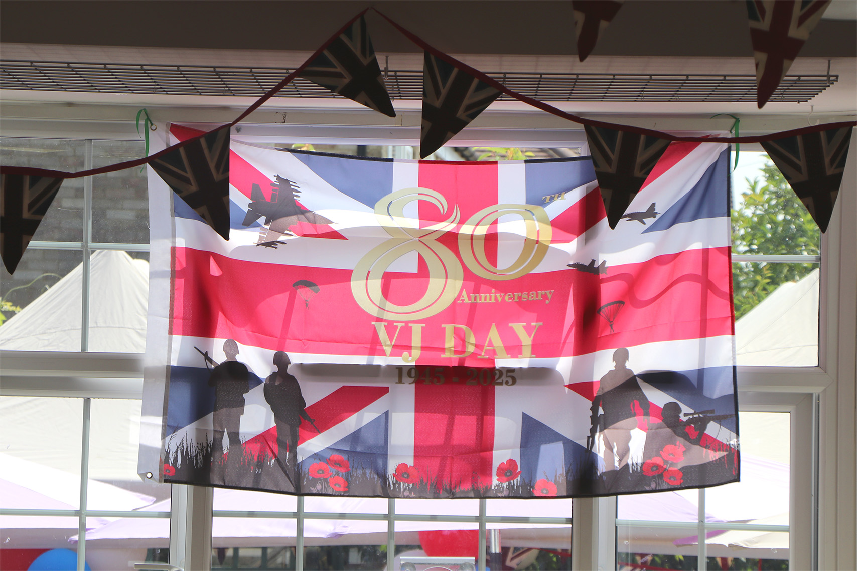 80th anniversary VJ Day banner featuring illustrations of planes and soldiers, in front of a window decorated with British flag bunting