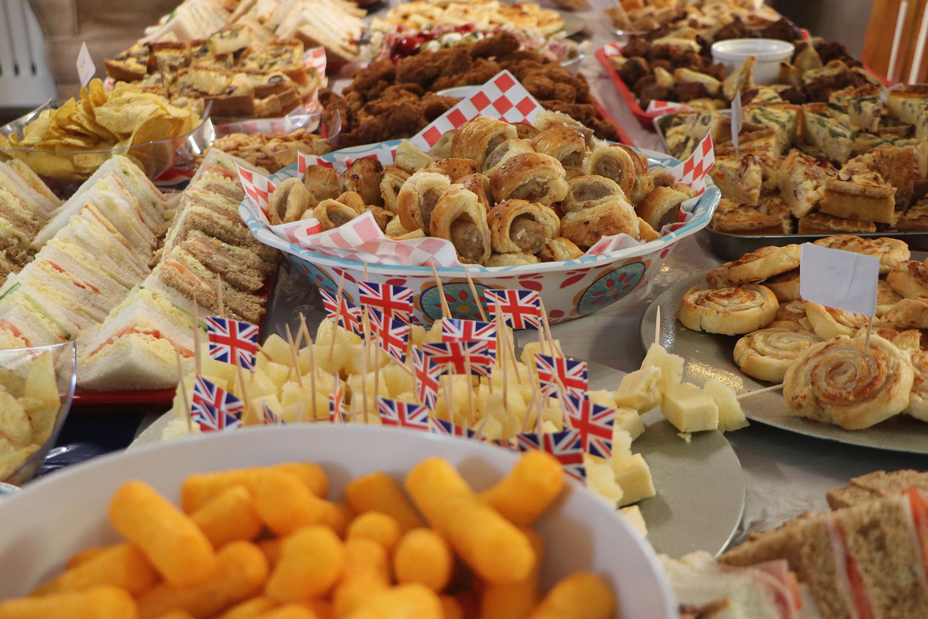 A buffet of food featuring sausage rolls, sandwiches, quiche, and samosas and cheese, decorated with British flags