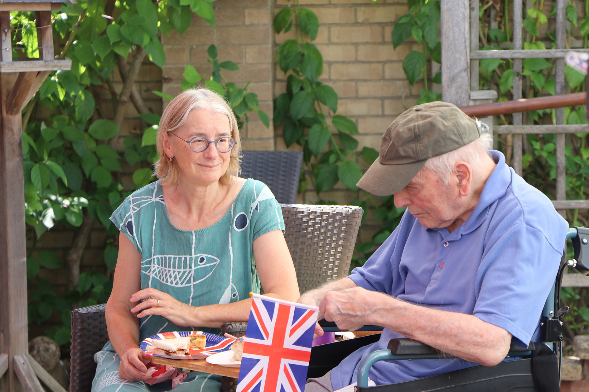 A resident holding a British flag and their family member sit in the care home garden and eat food