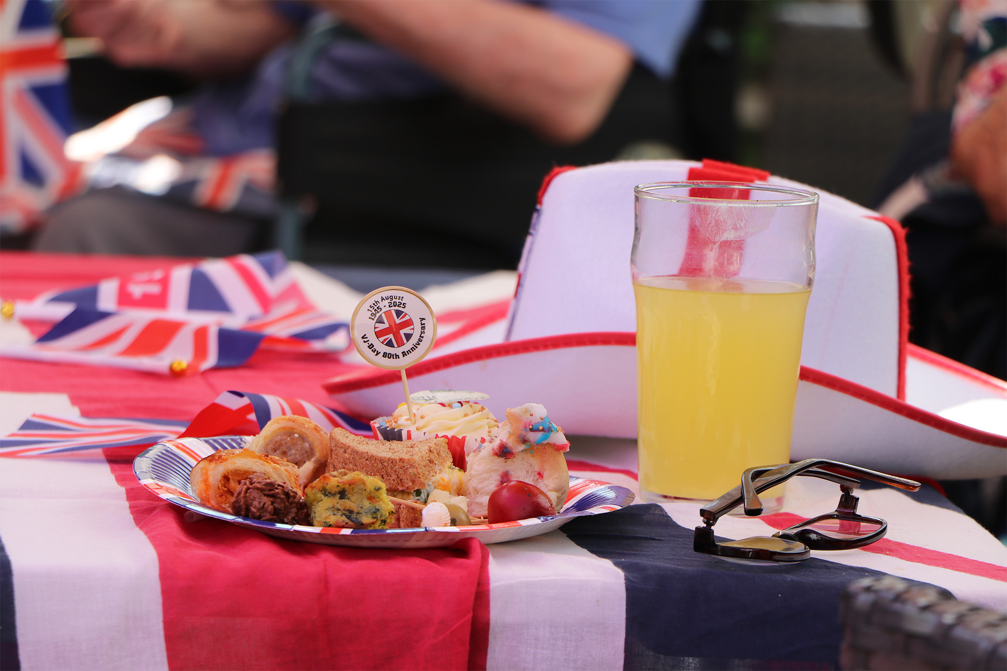 A plate of buffet food on a paper plate, next to a glass of orange squash, an England flag hat and a pair of glasses on a decorated table