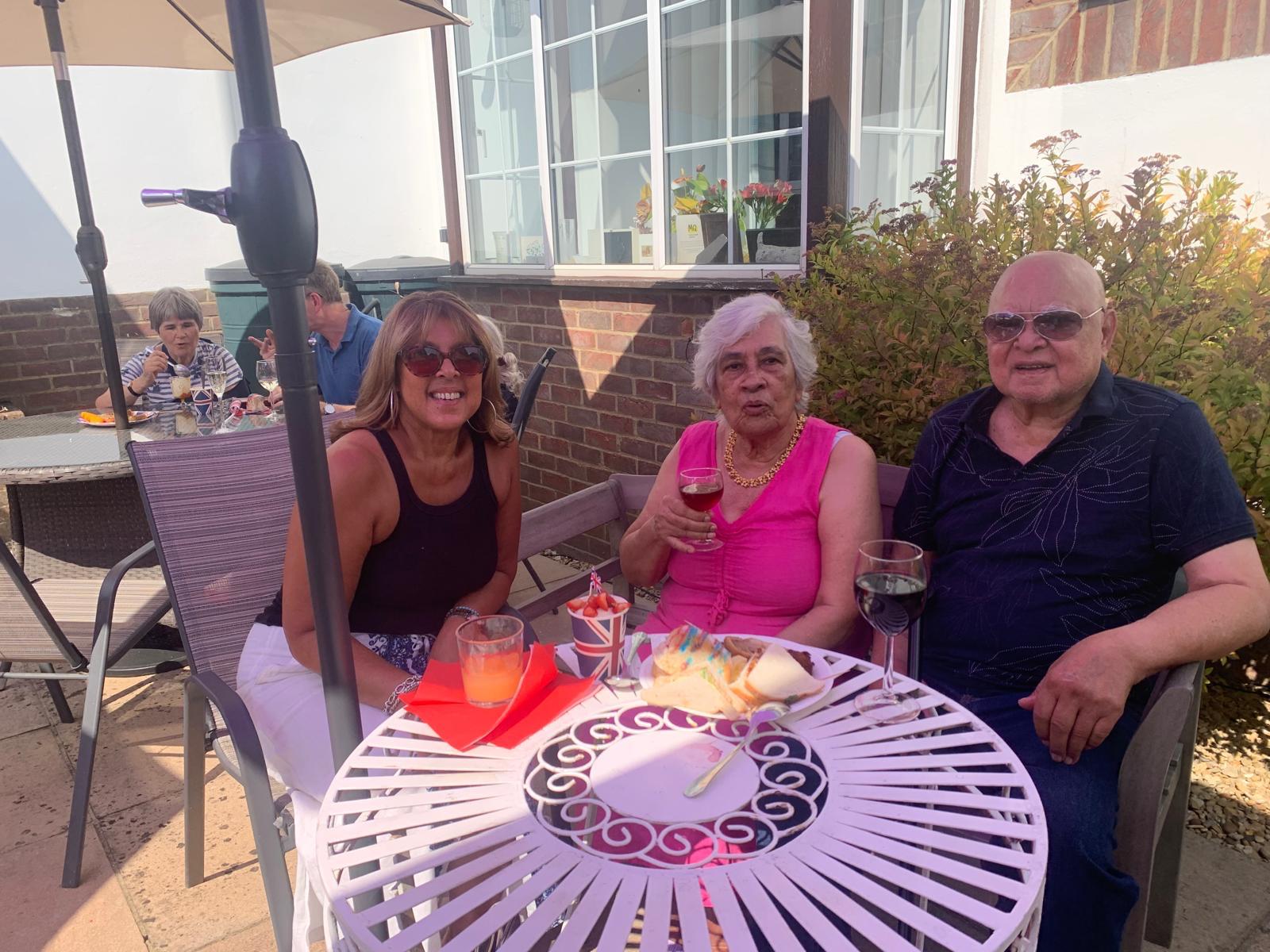 A resident and her 2 relatives smile, she wears a bright pink dress and holds a drink. In front of them is a plate of buffet food and strawberries and cream