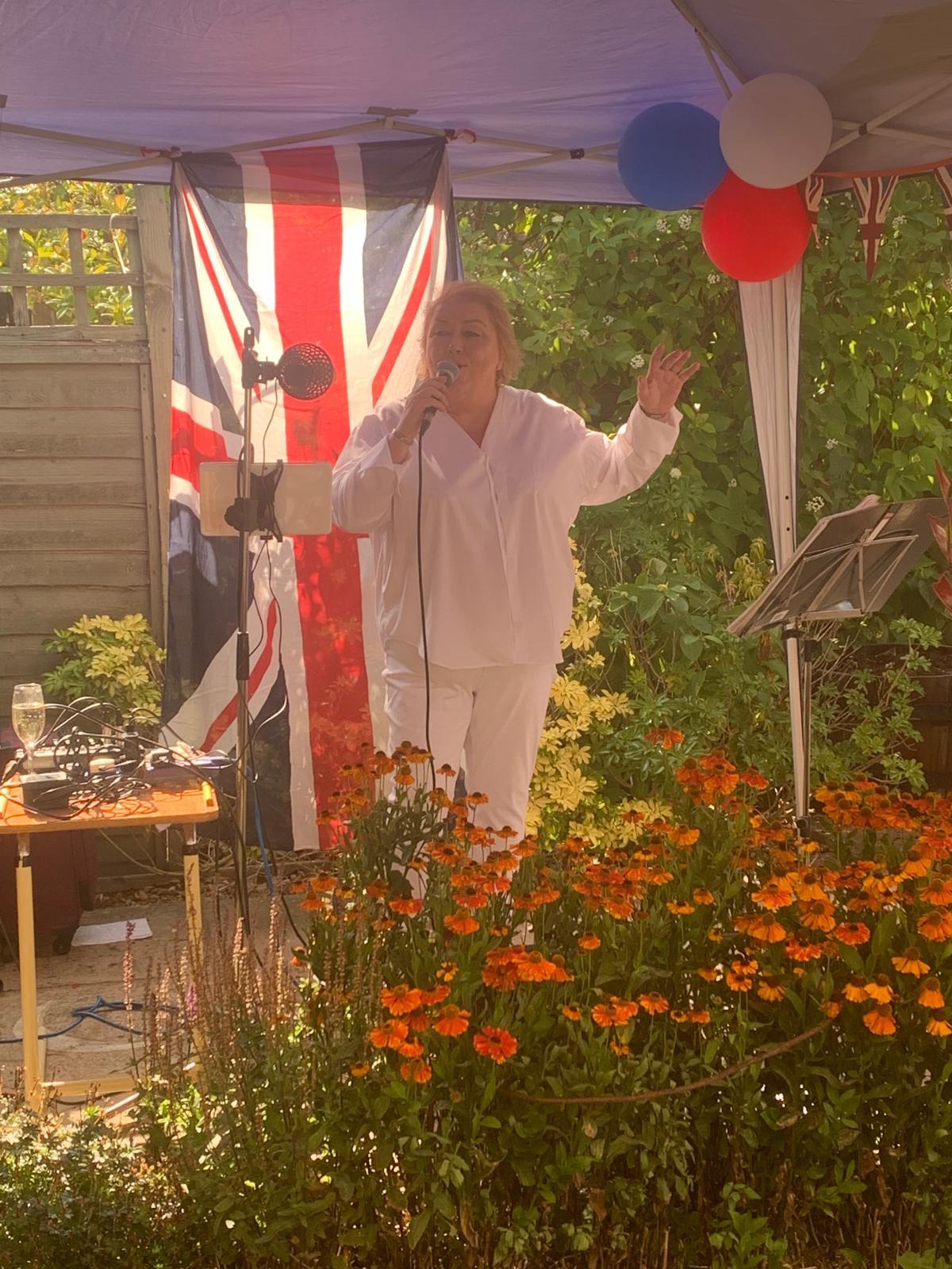 The singer wearing white stands under an awning in front of bright orange flowers, singing into the microphone, with the sun shining through a British flag behind her