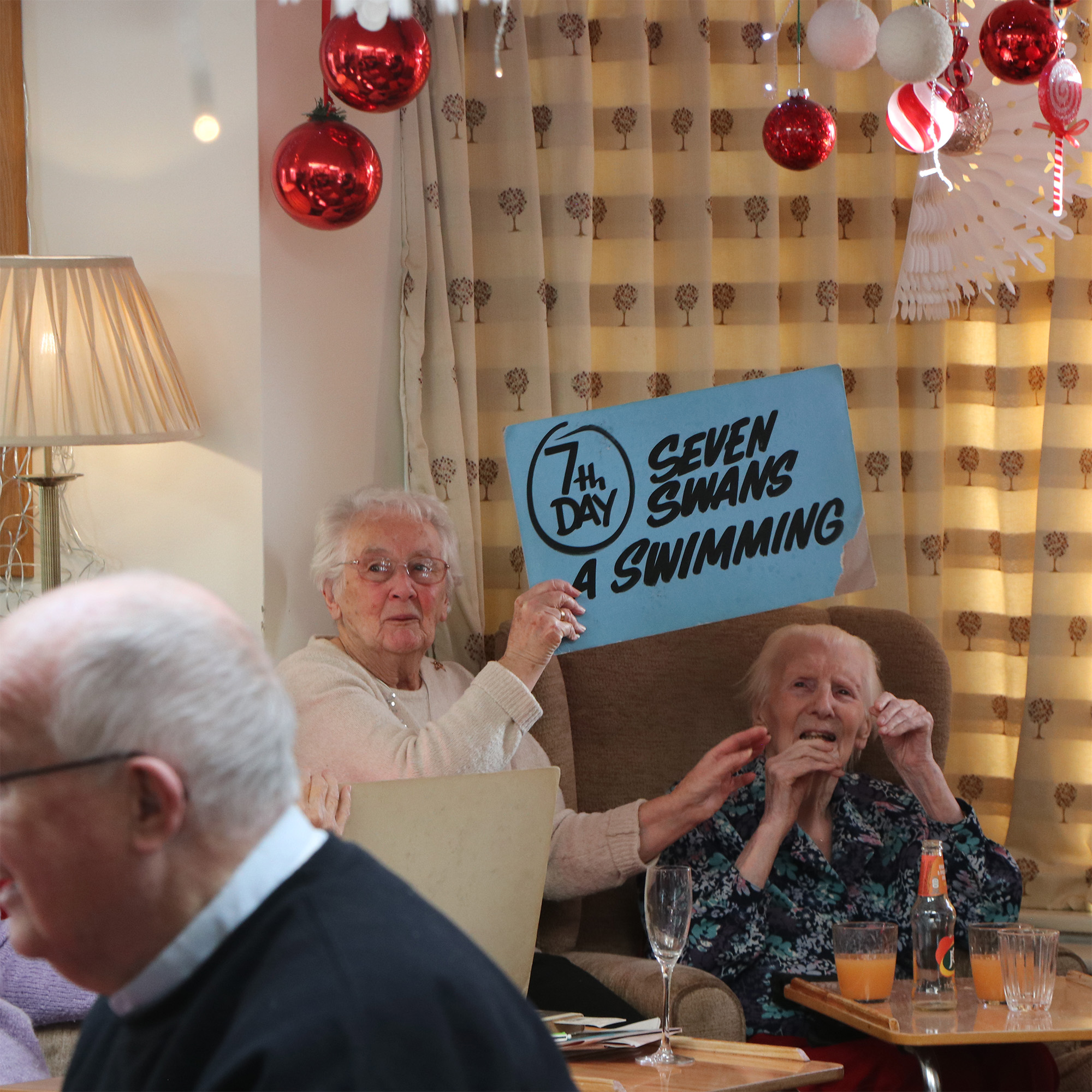 Two ladies taking part in a Christmas song game. One lady holds a large blue sign saying '7th Day' and 'Seven swans a swimming'.
