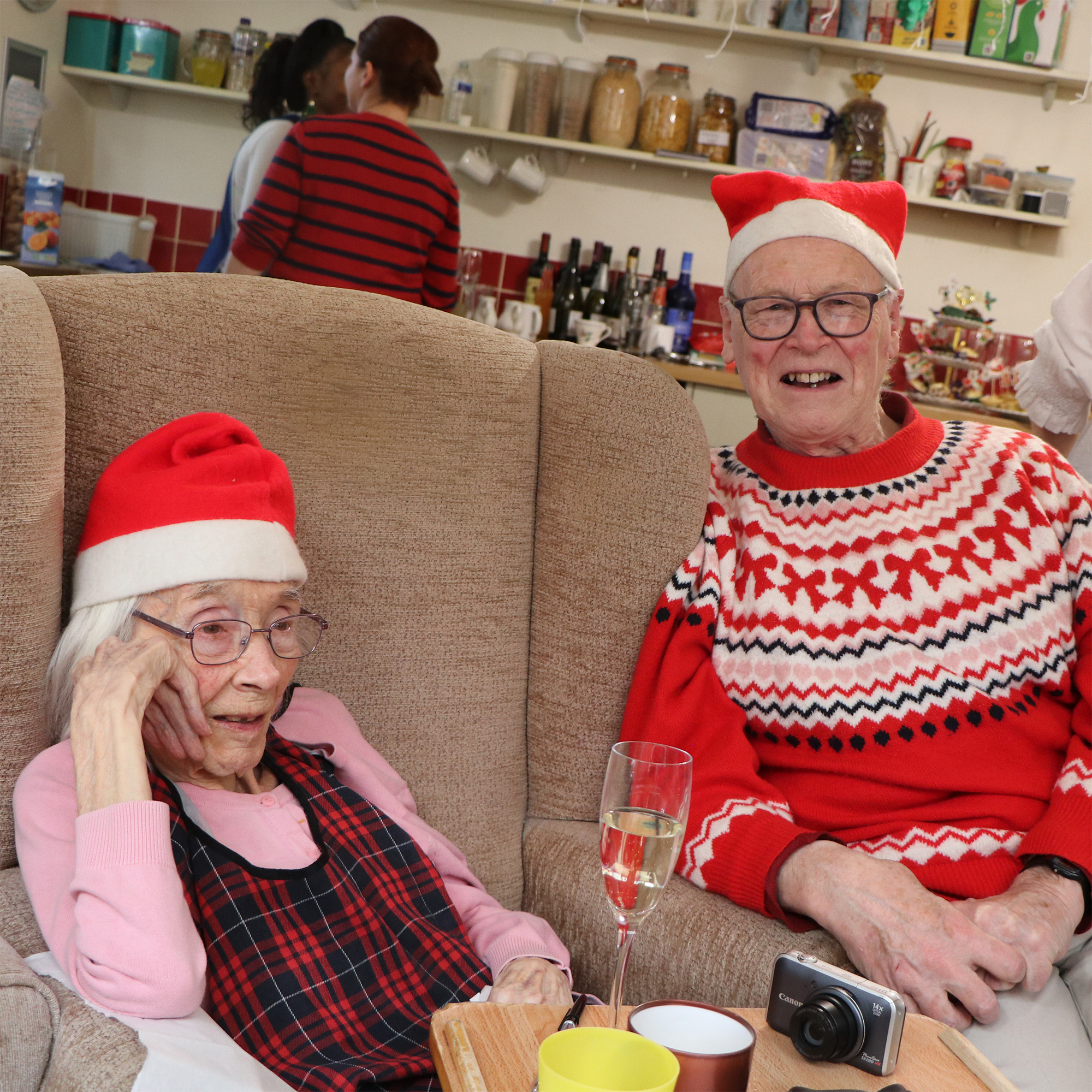 Two people wearing Santa hats smile while sitting in front of a flute of prosecco. The man is wearing a Christmas jumper with a red bow design. You can see staff members in the back chatting.