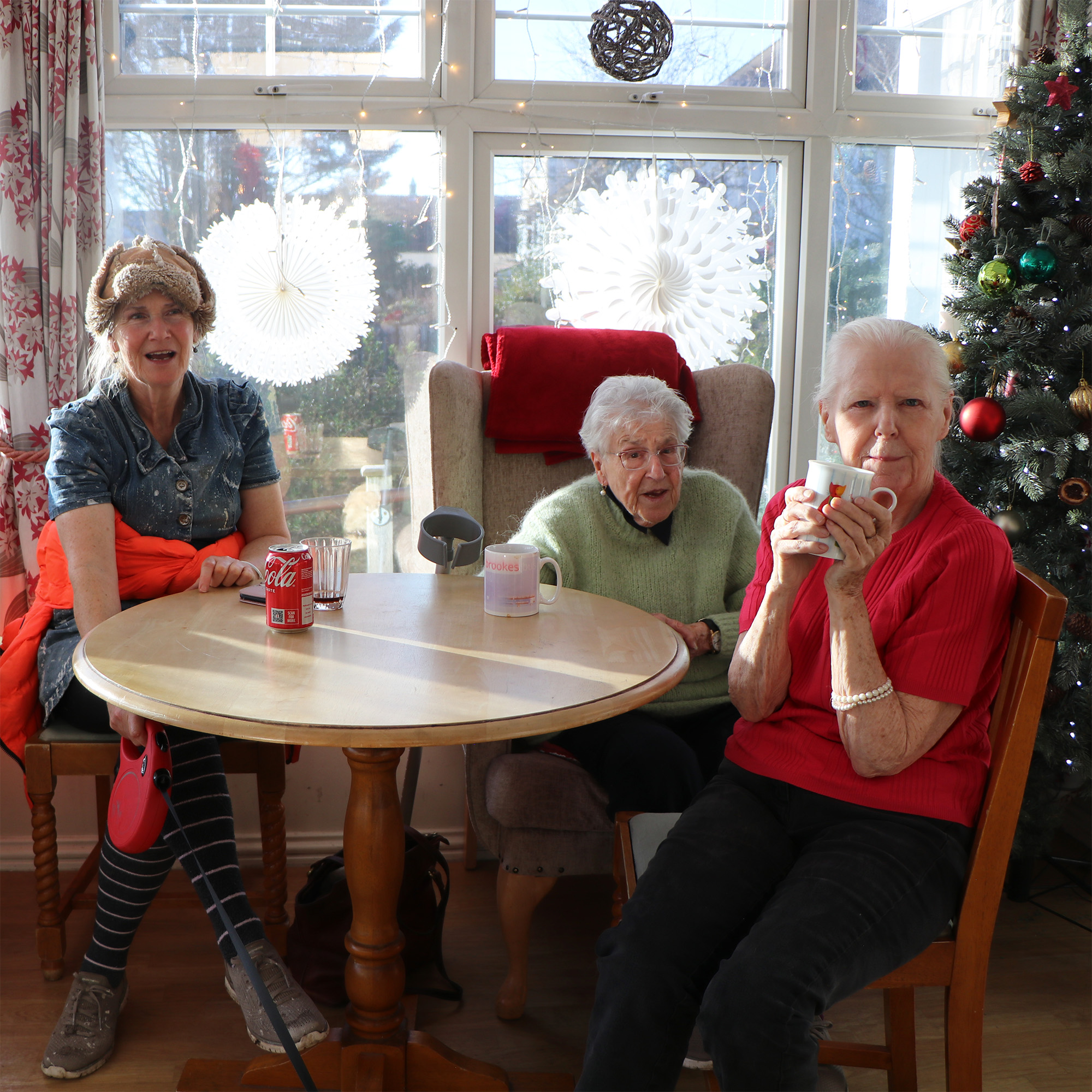 Three ladies sit around the table and smile while drinking cups of tea in-front of a Christmas tree and white snowflakes.