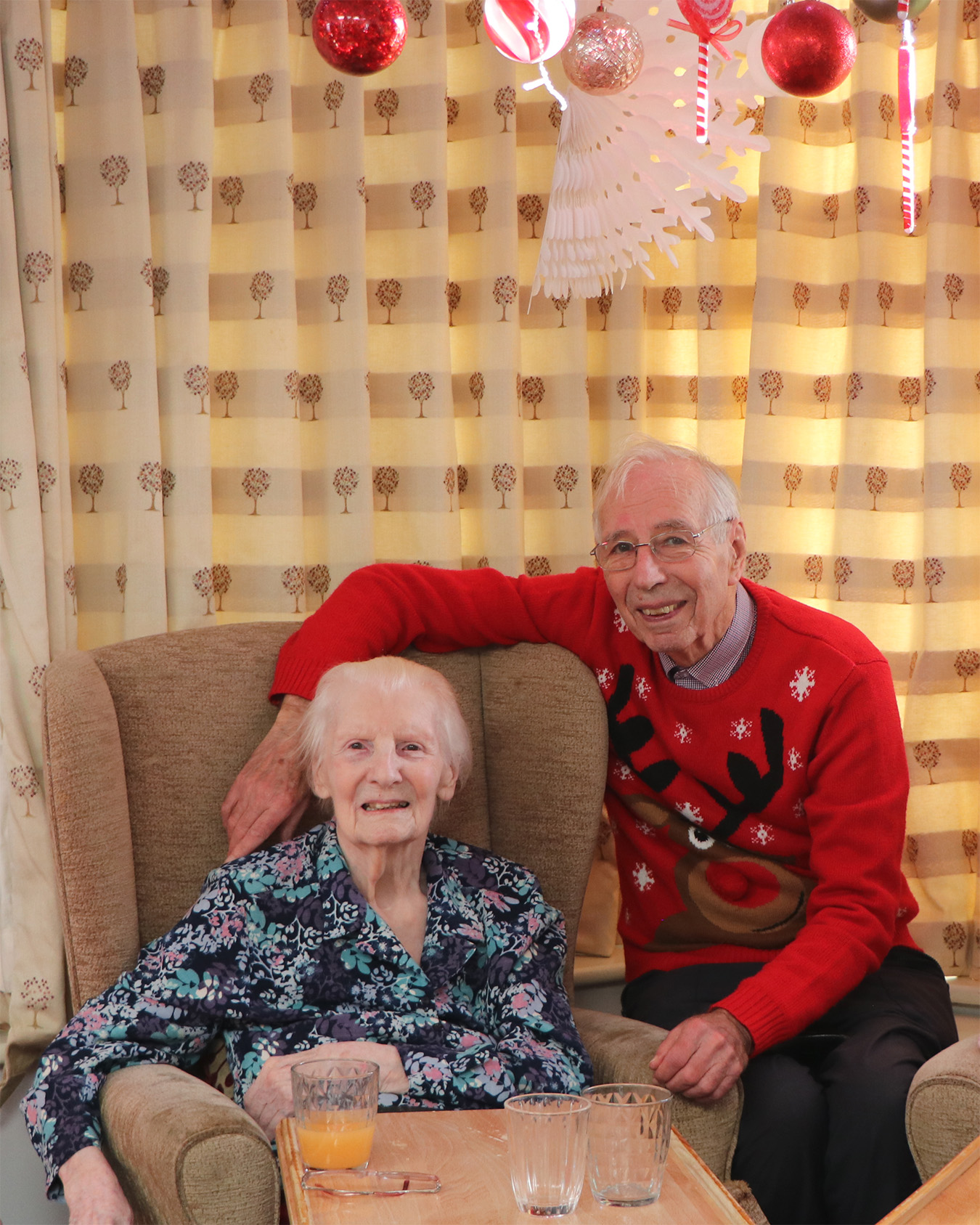 A resident sits in an armchair and smiles with a guest who is wearing a Christmas reindeer jumper.