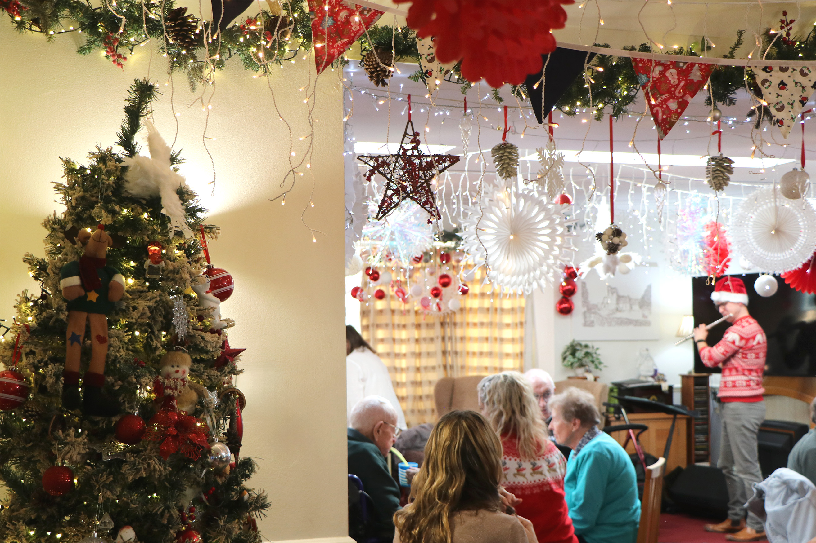 A lovely decorated Christmas tree is the focus of the image, while in the background the residents and their families are celebrating. There is also a musician playing the flute.