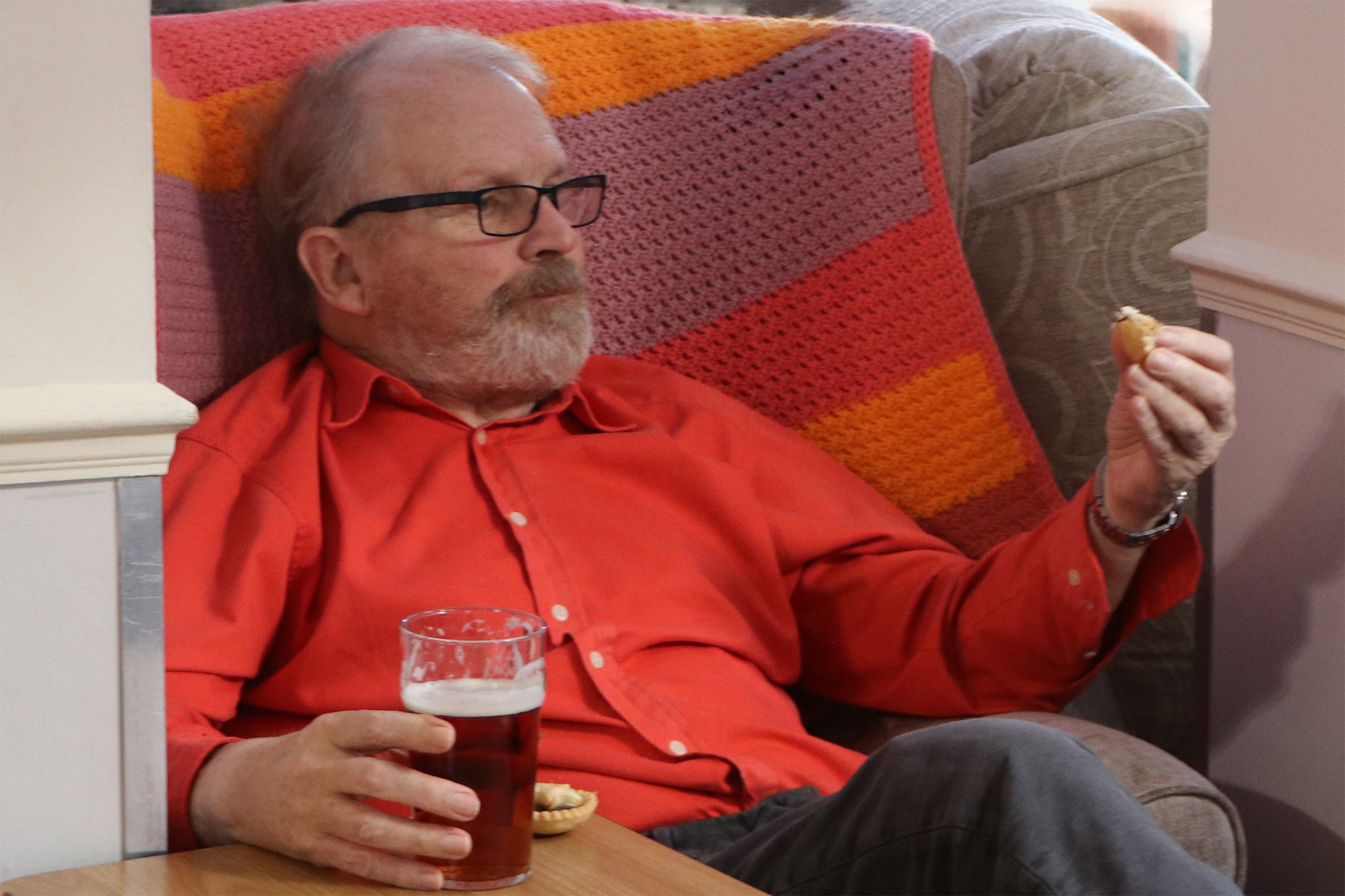 A man wearing a red shirt eats a mince pie and drinks a beer.