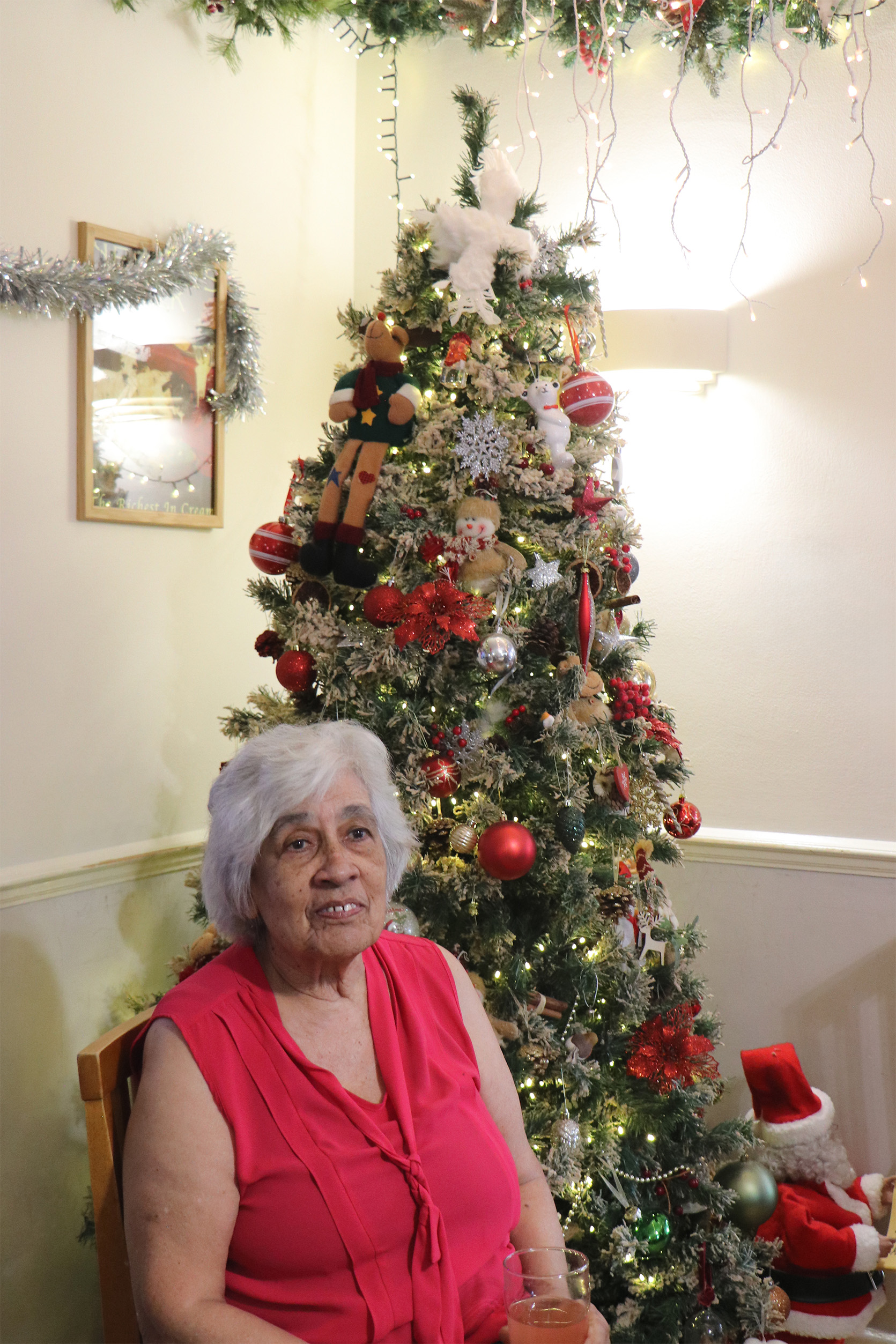 A lady wearing a red shirt smiles while she sits in front of a decorated Christmas tree.